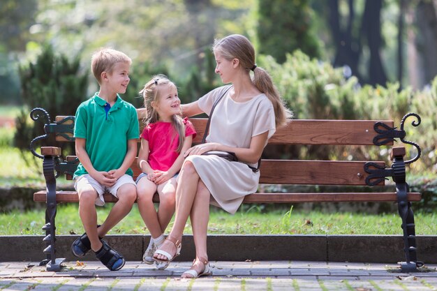 Young mother and two children boy and girl having fun time together on a bench in summer park.