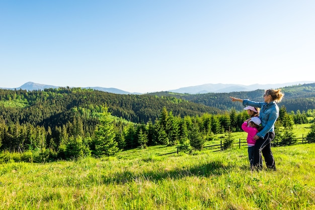Young mother and two little daughters travelers stand on a slope with a gorgeous view of the hills covered with dense fir forest against the blue sky on sunny warm summer day
