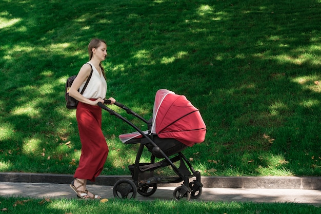 公園で乳母車を歩いて若い母親。赤ちゃんと一緒に新鮮な空気の中を歩きます。