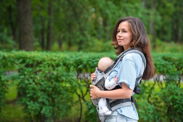 若い母親が緑の夏の公園でスリング バックパックで眠っている赤ちゃんと一緒に歩く