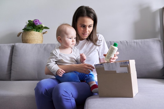 Young mother with baby in her arms unpacking parcel with cosmetics, household goods, sitting at home on the sofa. Online shopping, parcel delivery service