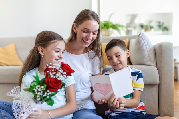 Young mother with a bouquet of roses laughs, hugging her son, and ?heerful girl with a card congratulates mom during holiday celebration in kitchen at home