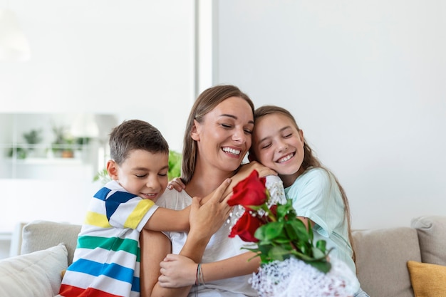 バラの花束を持った若い母親が笑い、息子を抱きしめ、カードを持った陽気な女の子が自宅のキッチンでの休日のお祝い中にお母さんを祝福します