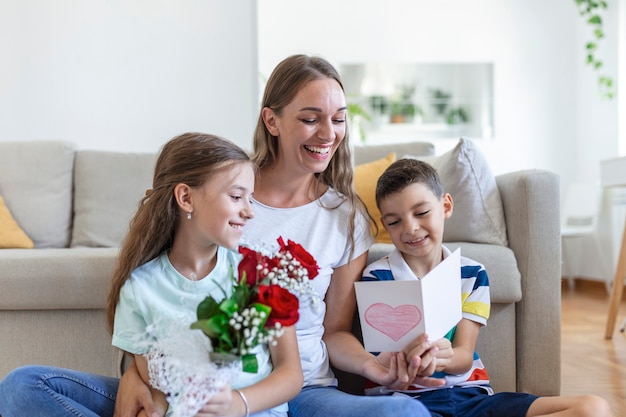 バラの花束を持った若い母親が息子を抱きしめて笑い、カードとバラを持った陽気な女の子が家での休日のお祝いの間にお母さんを祝福します。母の日