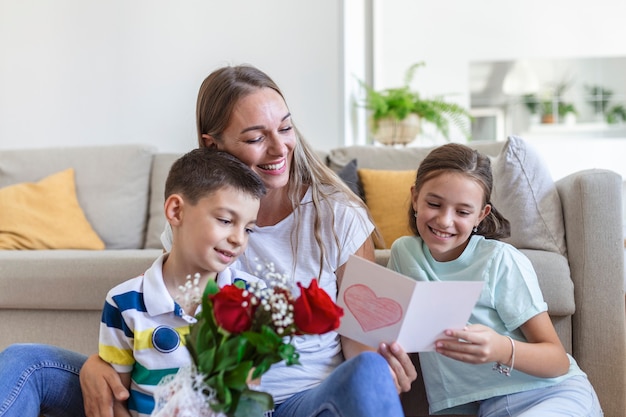 Young mother with a bouquet of roses laughs, hugging her son, and Ñheerful girl with a card and roses congratulates mom during holiday celebration in kitchen at home. mothers day