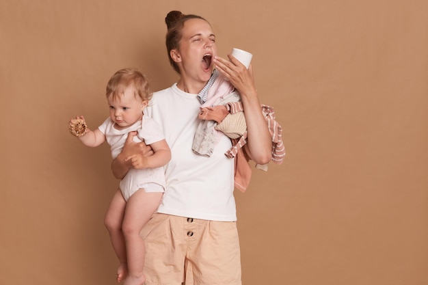 Young mother with dark hair wearing white t shirt standing with her baby daughter in hands yawning needs more sleep having sleepless nights during maternity leave isolated over brown background