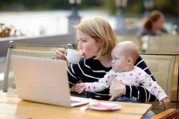 Young mother with her adorable baby girl working or studying on her laptop in outdoor cafe