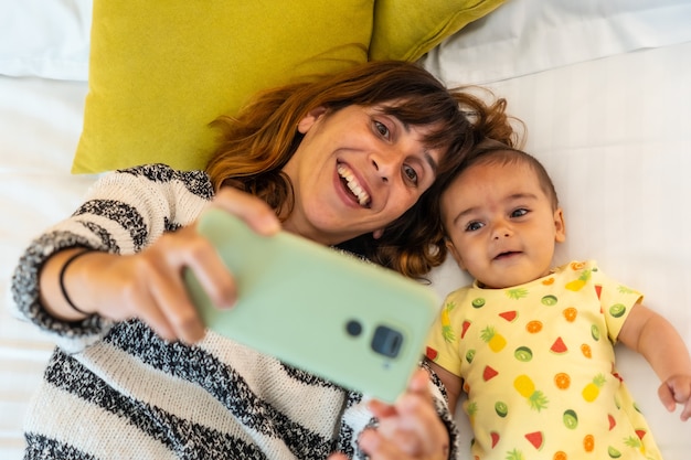 寝室のベッドで携帯電話を使って自分撮りをしている赤ちゃんを持つ若い母親、自宅の家族