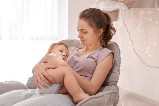 Young mother with her cute little baby sitting in armchair at home