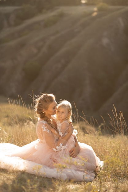 Young mother with a little daughter in pink dresses are sitting in the field, Mom hugs daughter, hugging her