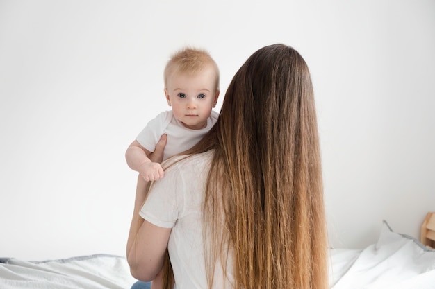 Young mother with long brown hair, sitting on bed holding cute little blond baby on white surface