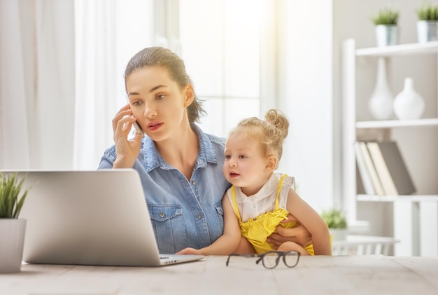 Молодая мать с малышом, работающим за компьютером, дома разговаривает по телефону.