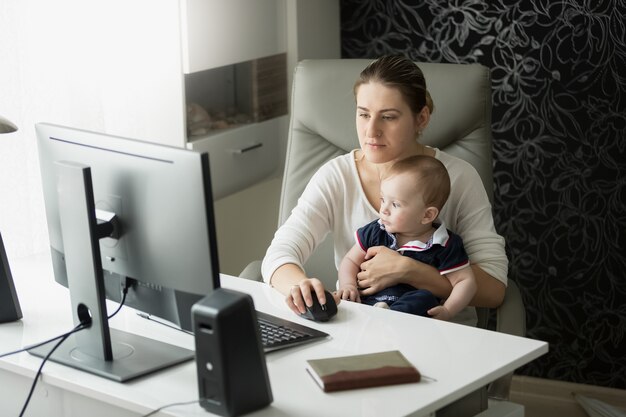 Young mother working at home office and taking care of her baby son