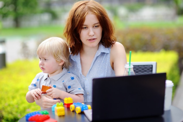 Young mother working oh her laptop and holding her sad toddler son