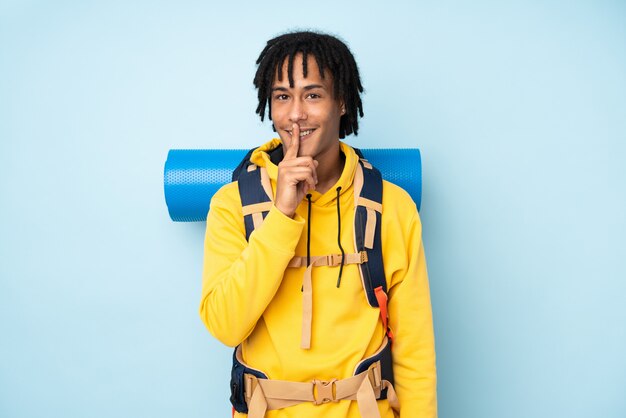 Young mountaineer african american man with a big backpack on a blue wall doing silence gesture