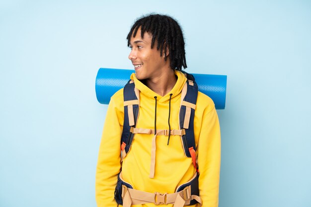 Young mountaineer african american man with a big backpack on a blue wall looking to the side