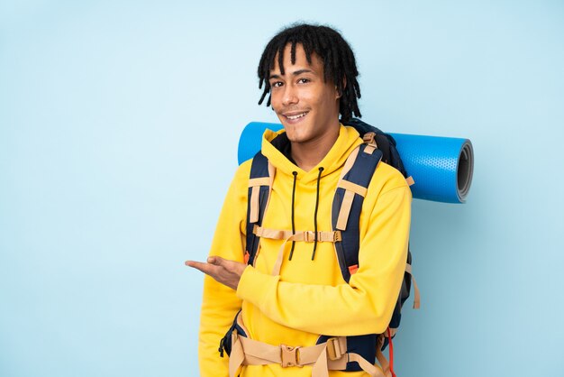 Young mountaineer african american man with a big backpack isolated on a blue background extending hands to the side for inviting to come