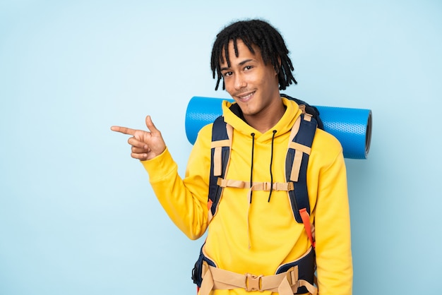 Молодой альпинист афроамериканец человек с большой рюкзак, изолированных на синем фоне, указывая пальцем в сторону