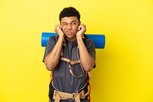 Young mountaineer African American man with a big backpack isolated on yellow background frustrated and covering ears