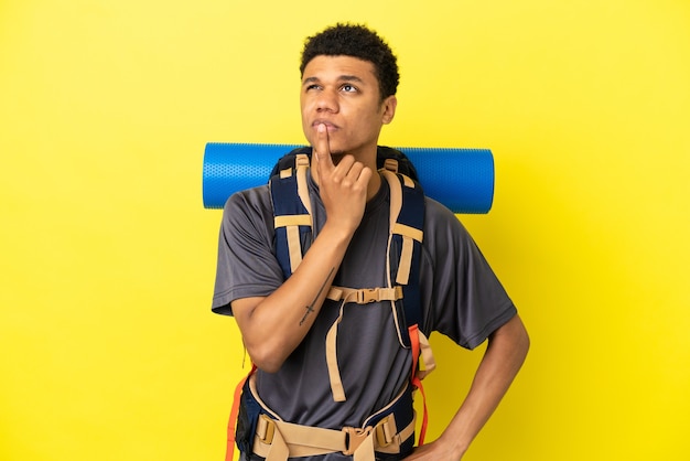 Young mountaineer African American man with a big backpack isolated on yellow background having doubts while looking up