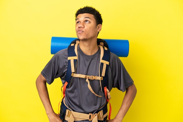 Young mountaineer African American man with a big backpack isolated on yellow background and looking up