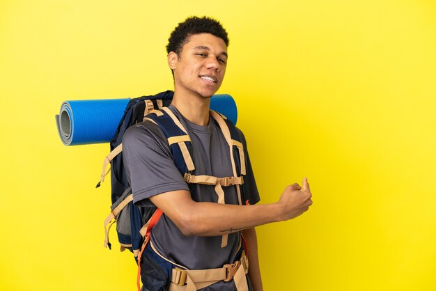 Young mountaineer African American man with a big backpack isolated on yellow background pointing back