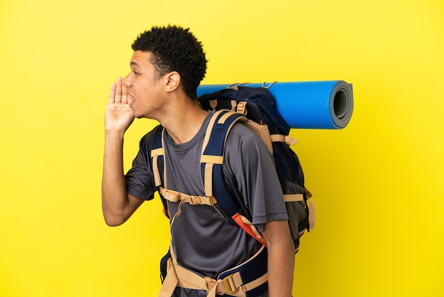 黄色の背景に分離された大きなバックパックを持つ若い登山家アフリカ系アメリカ人の男は、横に大きく開いた口で叫んでいます