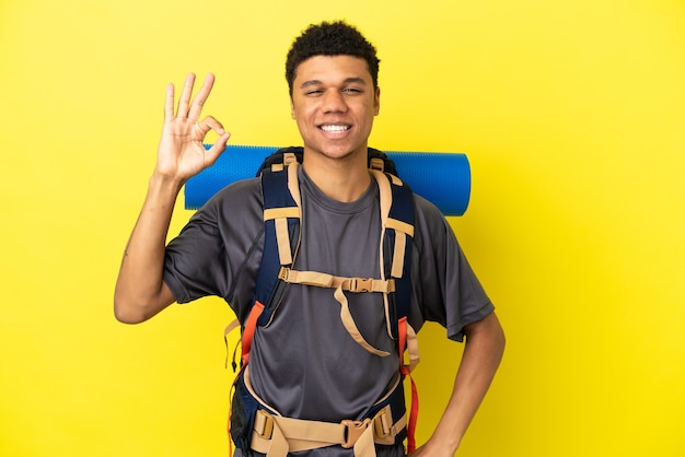指でOKサインを示す黄色の背景に分離された大きなバックパックを持つ若い登山家アフリカ系アメリカ人の男