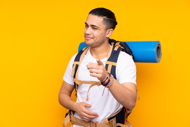 Young mountaineer asian man with a big backpack isolated on yellow points finger at you