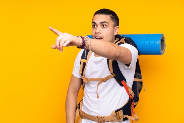 Young mountaineer asian man with a big backpack on yellow wall pointing away