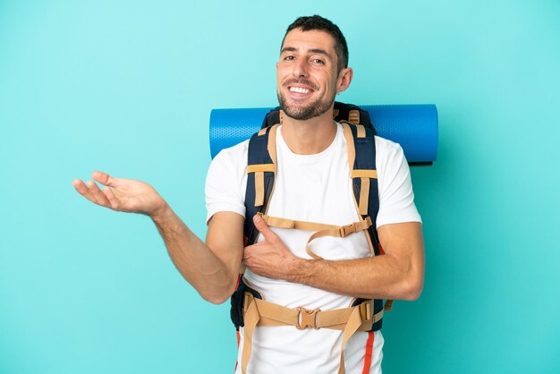 Young mountaineer caucasian man with a big backpack isolated on blue background extending hands to the side for inviting to come