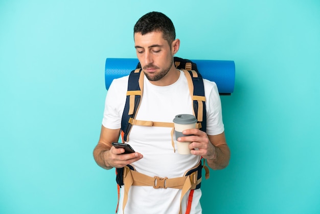 Young mountaineer caucasian man with a big backpack isolated on blue background holding coffee to take away and a mobile
