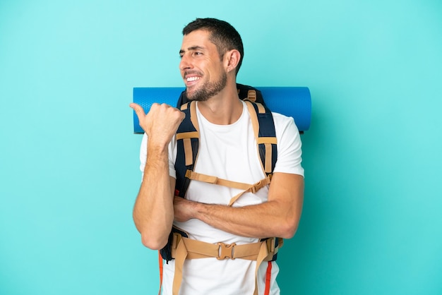 Young mountaineer caucasian man with a big backpack isolated on\
blue background pointing to the side to present a product