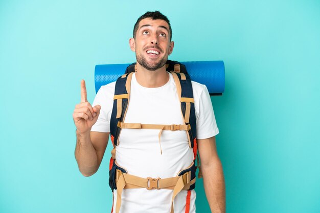 Young mountaineer caucasian man with a big backpack isolated on blue background pointing up and surprised