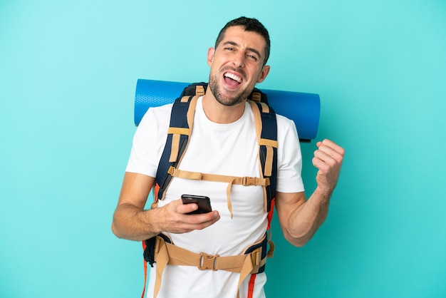 Young mountaineer caucasian man with a big backpack isolated on blue background with phone in victory position