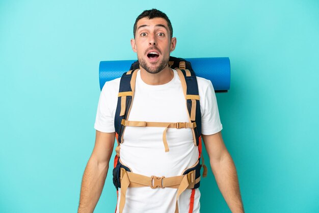 Young mountaineer caucasian man with a big backpack isolated on blue background with surprise facial expression