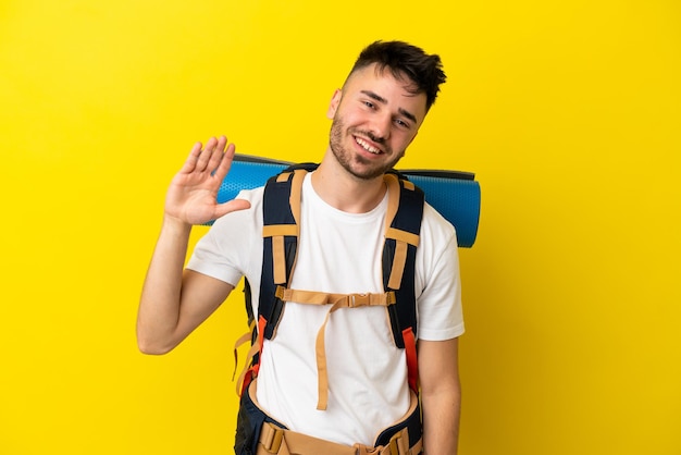 Young mountaineer caucasian man with a big backpack isolated on yellow background saluting with hand with happy expression