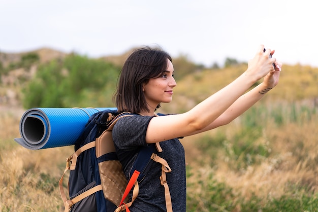 Young mountaineer girl taking a selfie with the mobile with a big backpack at outdoors