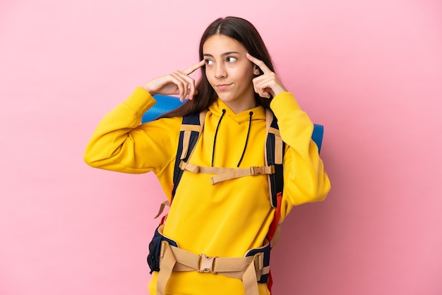 Young mountaineer girl with a big backpack isolated on pink background having doubts and thinking