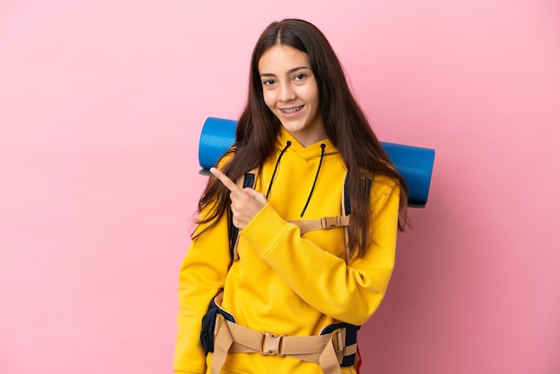 Young mountaineer girl with a big backpack isolated on pink background pointing to the side to present a product