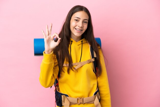 Young mountaineer girl with a big backpack isolated on pink background showing ok sign with fingers