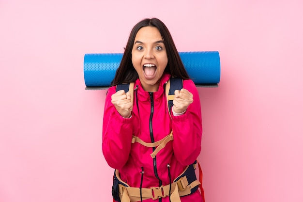 Young mountaineer girl with a big backpack over isolated pink wall celebrating a victory in winner position