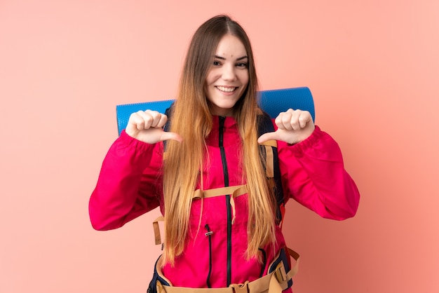 Young mountaineer girl with a big backpack isolated on pink wall proud and self-satisfied