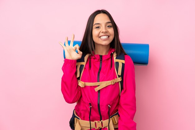 Молодая девушка альпиниста с большим рюкзаком на розовой стене, показывая знак ОК с пальцами