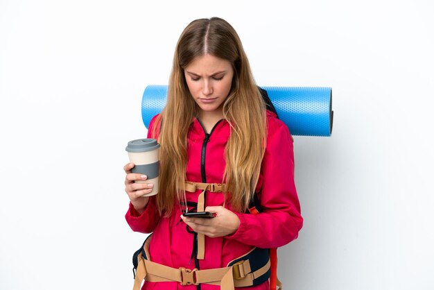 Молодая альпинистка с большим рюкзаком на изолированном белом фоне держит кофе на вынос и мобильный телефон
