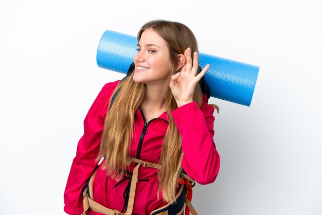 Young mountaineer girl with a big backpack over isolated white background listening to something by putting hand on the ear