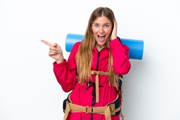 孤立した白い背景の上に大きなバックパックを持った若い登山家の女の子は驚いて、人差し指を横に向けています