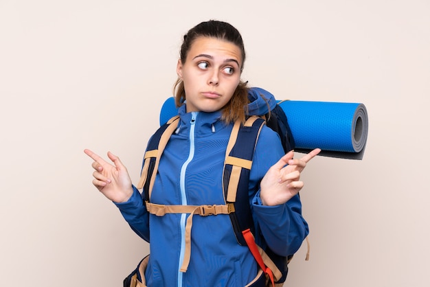 Young mountaineer girl with a big backpack pointing to the laterals having doubts