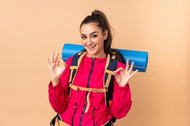 Young mountaineer girl with a big backpacker isolated on beige showing an ok sign with fingers