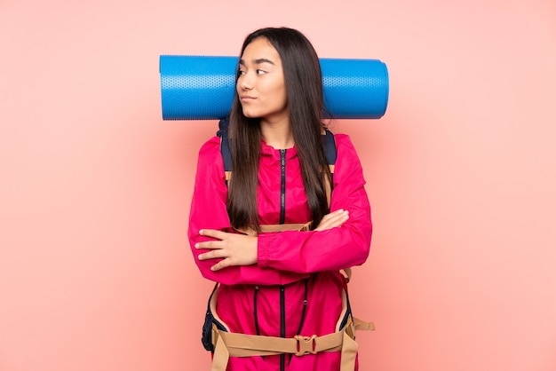 Young mountaineer Indian girl with a big backpack isolated on pink background looking side